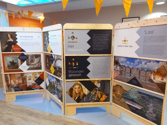 Informatiepaneel over Paleis Het Loo met foto's van historische portretten, paleisinterieurs, tuinen en het museum, opgesteld in een binnenruimte met houten presentatiewanden en oranje vlaggetjes.