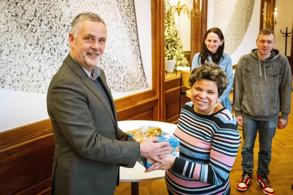 Vier personen in een representatieve ruimte van Paleis Het Loo. Op de voorgrond overhandigt een persoon een ingepakt cadeau aan een andere persoon. Op de achtergrond staan twee andere personen. De ruimte heeft houten wandpanelen, spiegels, kroonluchters en een kerstboom.