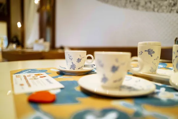 Koffiekopjes, schoteltjes en een suikerpot met blauw bloemendecor staan op een tafel met een kleurrijk patroon. Op de voorgrond ligt een informatiekaart. De foto is genomen in een ruimte van Paleis Het Loo met houten wandpanelen en spiegels op de achtergrond.