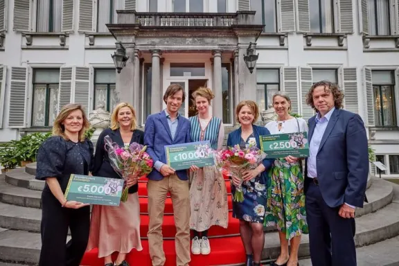 Zeven personen staan op een rode loper voor een historisch gebouw en houden cheques en boeketten bloemen vast. Op de cheques staan bedragen van €2.500, €5.000 en €10.000 vermeld als onderdeel van de VriendenLoterij Kasteelprijs.
