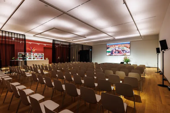 Evenementruimte in Paleis Het Loo met rijen stoelen gericht op een presentatiescherm, zwarte transparante gordijnen, houten vloer en een verlichte wand met vitrines.