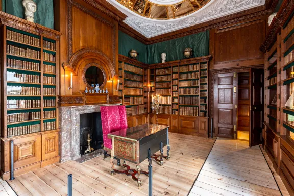 Bibliotheek met boekenkasten vol oude boeken, een rode leesstoel, een tafeltje, een wit plafond met spiegels en een houten vloer