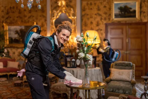 Een medewerker museale schoonmaak lacht terwijl ze een tafel in het paleis schoonmaakt