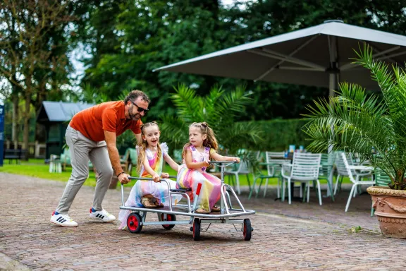 Man duwt speelauto met twee kinderen in prinsessenjurken erin