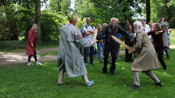 Groep mensen in een parkachtige omgeving tijdens een buitentheater- of spelactiviteit. Twee deelnemers staan tegenover elkaar met grote kartonnen zwaarden, terwijl omstanders in een halve cirkel toekijken. Eén deelnemer draagt een kartonnen kroon. Op de achtergrond zijn bomen, gras en wandelpaden zichtbaar.