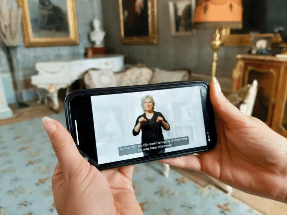 Een bezoeker houdt een mobiel vast met daarop de NGT video tour van het Huis van Wilhelmina