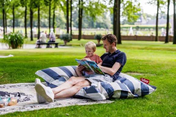 Man leest kind voor op groot blauw-wit gestreept kussen in de tuinen