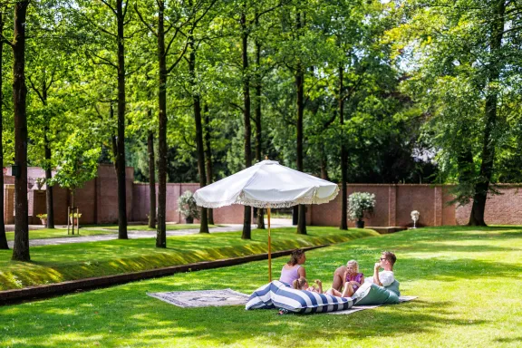Gezelschap ontspant op grote kussens en een kleed onder een witte parasol in een groene tuin van Paleis Het Loo. Op de achtergrond staan hoge bomen, een gazon en bakstenen muren die de tuin omringen.