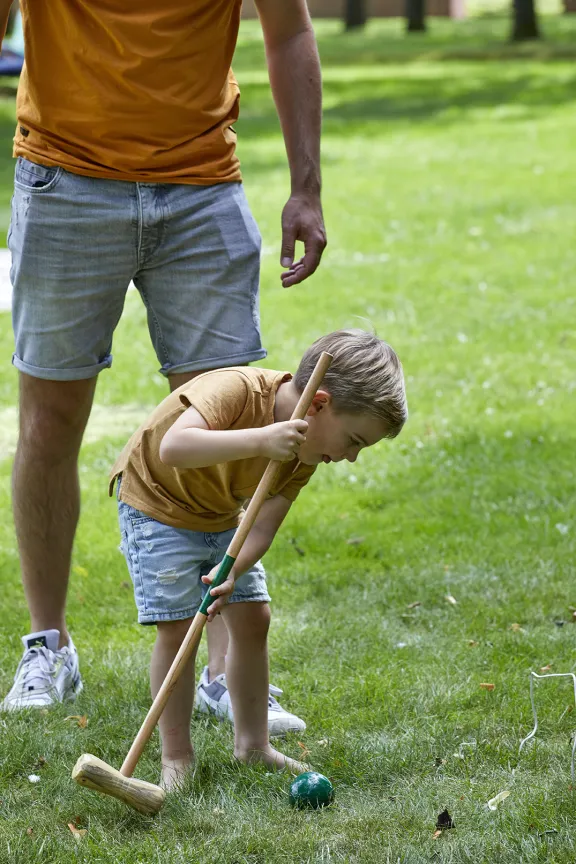 Een jongen met een croquet hamer slaat een bal met zijn vader en de chillweide op de achtergrond