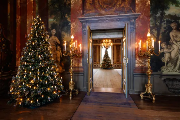 Links op de houten vloer staat een grote, rijkelijk verlichte kerstboom met traditionele kaarsjes en zilveren decoraties. In het midden sieren twee grote, gouden kandelaars met brandende kaarsen een geopende dubbele deur. Door de deuropening is in de aangrenzende kamer een tweede, verlichte kerstboom te zien onder een kristallen kroonluchter. De wanden van de zaal zijn bedekt met klassieke, barokke muurschilderingen van weidse landschappen, geflankeerd door marmeren sculpturen.
