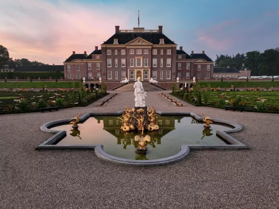 Ein großes Gebäude mit formellen Gärten und einem Brunnen im Vordergrund. Die Szene scheint bei Sonnenaufgang oder Sonnenuntergang zu sein, mit einem weichen, farbenfrohen Himmel.