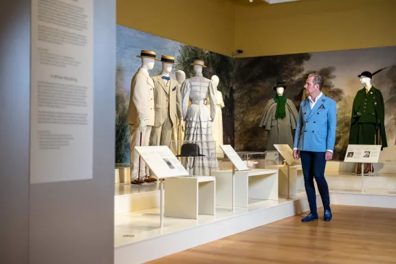 Een bezoeker in een blauw jasje, zwarte pantalon en herenschoenen loopt langs verschillende buitenkleding kostuums. Pakken van mannen in wit en beige met zonnehoeden, witte japons en ook kostuums in verschillende kleuren groen