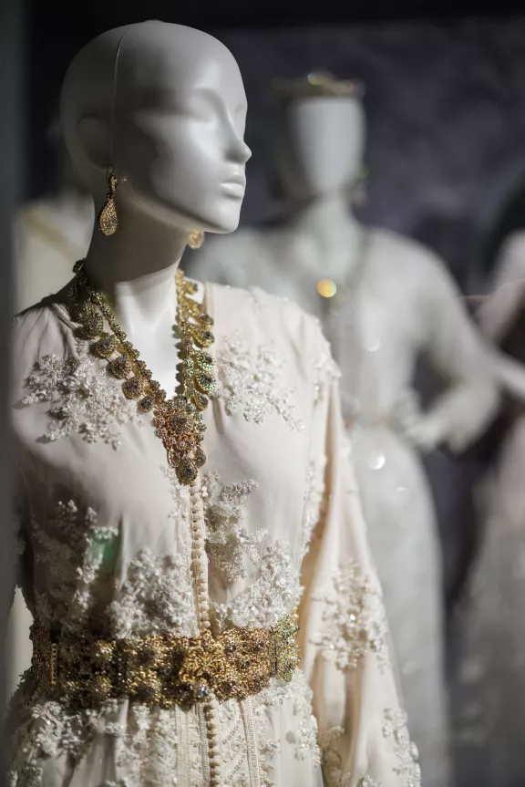 Een mannequin met een witte jurk met gouden en witte details en gouden oorbellen onderdeel van het gedeelte rouw en trouw van de tentoonstelling Dresscodes