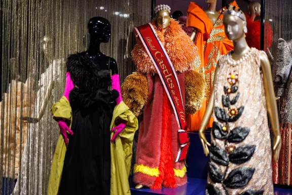 Links (zwart met geel/groen): Een elegant ensemble van de Belgische ontwerper Dries van Noten. Het bestaat uit een zwarte jurk gecombineerd met een opvallende geelgroene jas en knalroze handschoenen. Midden (oranje/rood met sjerp): De "Size Queen"-jas/jurk van het Nederlandse modeduo Viktor & Rolf. Dit extravagante ontwerp met extreem volumineuze kragen en mouwen is voorzien van een sjerp met de tekst "Build Castles in the Air". Rechtsvoor (goud/beige met bladeren): Een adembenemende, rijk gedecoreerde jurk van de Chinese couturier Guo Pei. Deze creatie valt op door het uiterst gedetailleerde handgemaakte borduurwerk met zilverdraad en glanzende stenen kralen.
