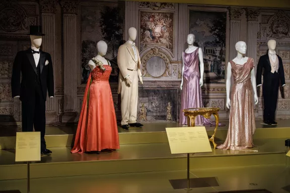 Verschillende gala kostuums. Van links naar rechts: een zwarte tuxedo, een 1960 avondjurk van ontwerper Ann Lowe, gekenmerkt door een levendige koraalrode kleur en gedetailleerde bloemversieringen op de schouder, een crèmekleurige rokkostuum uit de Biedermeier-periode, een paarse avondjapon met watervalhals, een roségouden avondjurk en een rokkostuum met een witte vlinderdas, vest en pantalon.
