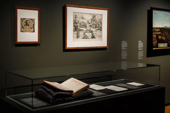 Twee schilderijen en prenten op de muur met een vitrine met daarin een opengeslagen boek en brieven