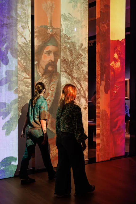mensen lopen langs lamellen met projectie van schilderij met ottomaanse figuren
