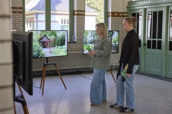 Twee vrouwelijke bezoekers kijken in het stallenplein naar schermen met beelden van het kunstwerk Searching for Lost Seeds
