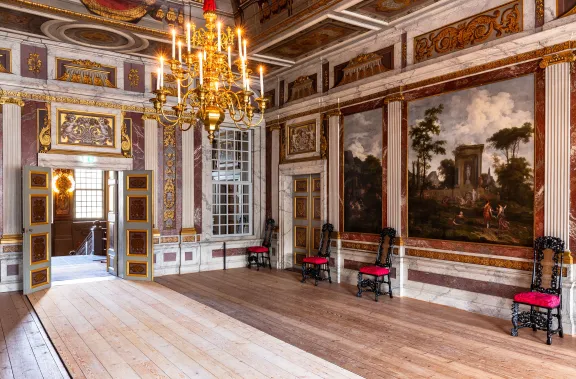 De grote zaal met bruine stoelen met rode kussens, een kroonluchter en muren en plafond versierd en geschilderd in barok stijl