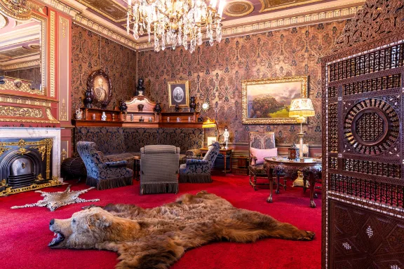De zitkamer van prins Hendrik met een rode vloer met een berenvel en katachtig vel op de grond, donkergrijze stoelen en bank, een open haard, schilderijen, lampen en een kroonluchter