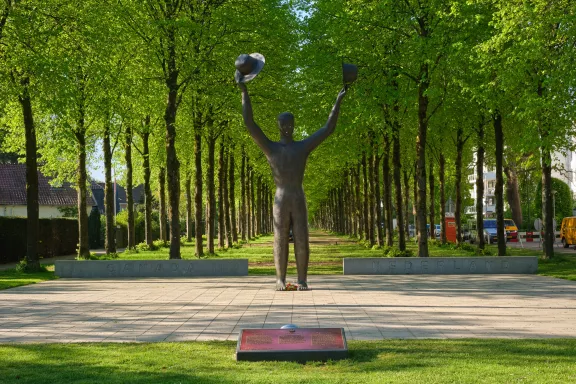 Een symmetrische foto van het bronzen standbeeld 'De Man met Twee Hoeden' op een stenen plein, omringd door een weelderige laan met groene bomen in Apeldoorn.