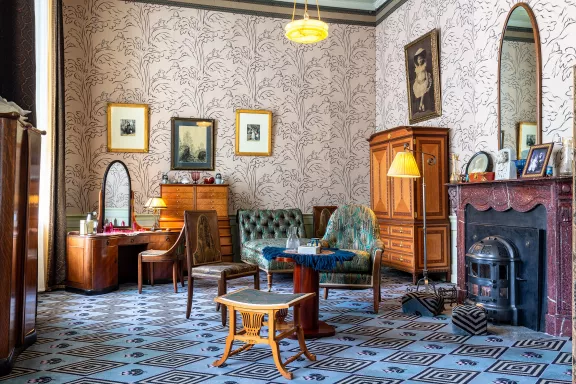 Een historisch interieur van een koninklijke zit- en slaapkamer in Paleis Het Loo, ingericht in de stijl van 1927. De kamer heeft druk behang met beige bladmotieven en een blauw tapijt met een geometrisch ruitpatroon. In de ruimte staan houten meubels, waaronder een kaptafel met spiegel, een ladekast en een houten kledingkast. Centraal staan een groen gecapitonneerde bank en fauteuils rond een kleine ronde tafel. Rechts bevindt zich een donkerrode marmeren schouw met een ingebouwde gietijzeren kachel, met daarboven een hoge, smalle boogspiegel.