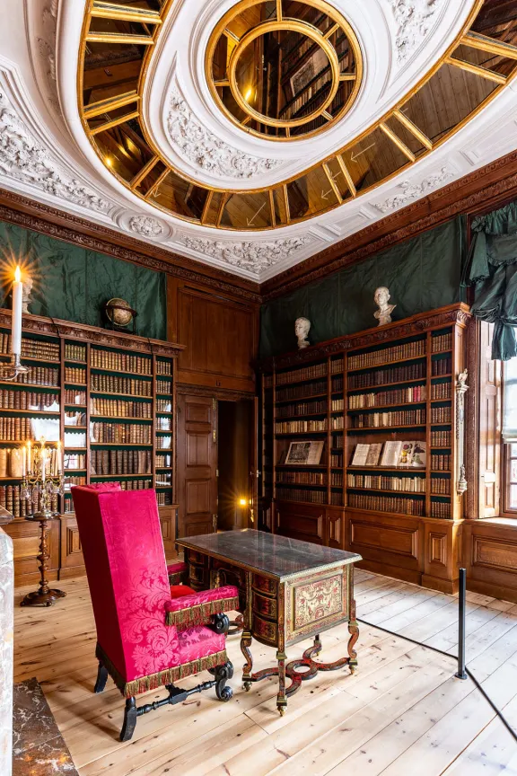 Bibliotheek met boekenkasten vol oude boeken, een rode leesstoel, een tafeltje, een wit plafond met spiegels en een houten vloer