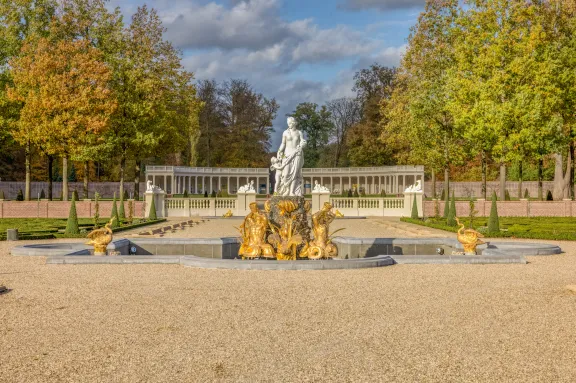 Een symmetrische foto van de Venusfontein in de baroktuinen van Paleis Het Loo in Apeldoorn. De fontein heeft een centrale, witte marmeren sculptuur van de godin Venus, met daaronder glanzende, gouden beelden van zeemeermannen waar water uit stroomt. De fontein staat in een grote, halfronde vijver op een uitgestrekt grindplein. Op de achtergrond is een lange, witte zuilengalerij (colonnade) te zien, geflankeerd door symmetrisch gesnoeide hagen en bomen in herfstkleuren onder een lichtbewolkte blauwe lucht.